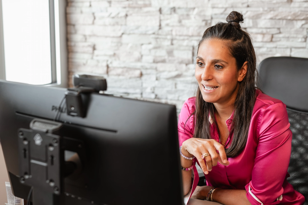 Eine lächelnde Frau sitzt am Schreibtisch vor einem großen Monitor und gestikuliert beim Arbeiten am Computer.